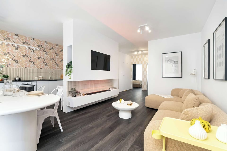 An open-concept living space connects the lounge area with a dining table and chairs in the background. A flat-screen television is mounted on the wall above a stylish white fireplace console.