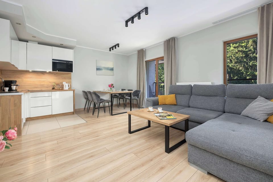 A spacious open-plan living room connected to a modern kitchenette. Large windows provide plenty of daylight and views of the greenery.