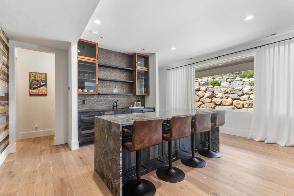 Gather around the stylish bar seating with rustic leather stools, perfect for morning coffee or evening cocktails. Natural light floods the space through flowing curtains, revealing a serene stone wall view outside.