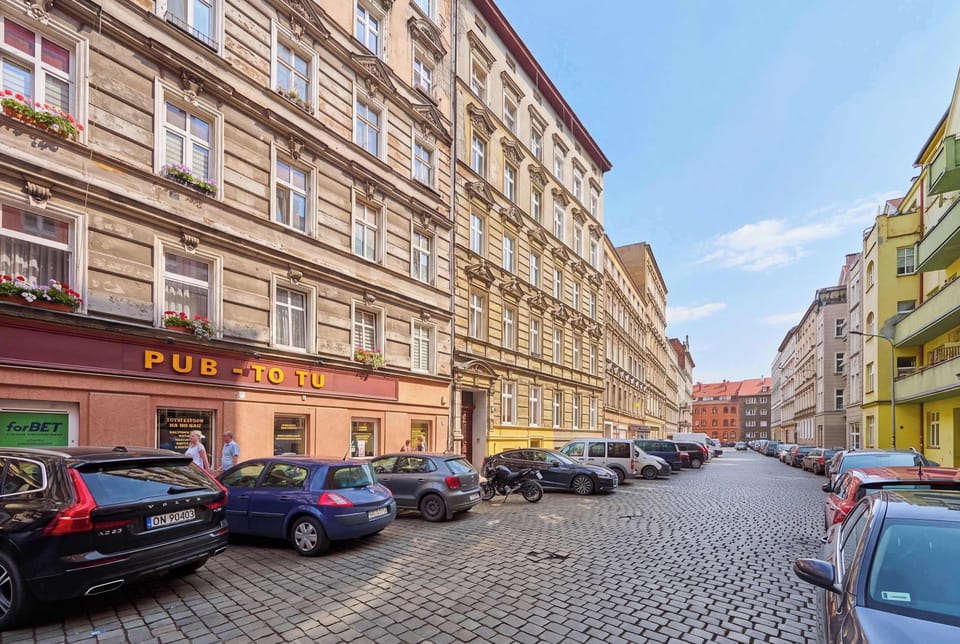 A wide street-level view highlights the beautiful historic architecture of the neighborhood's apartment buildings. The cobblestone road and rows of parked vehicles emphasize the charming urban setting.