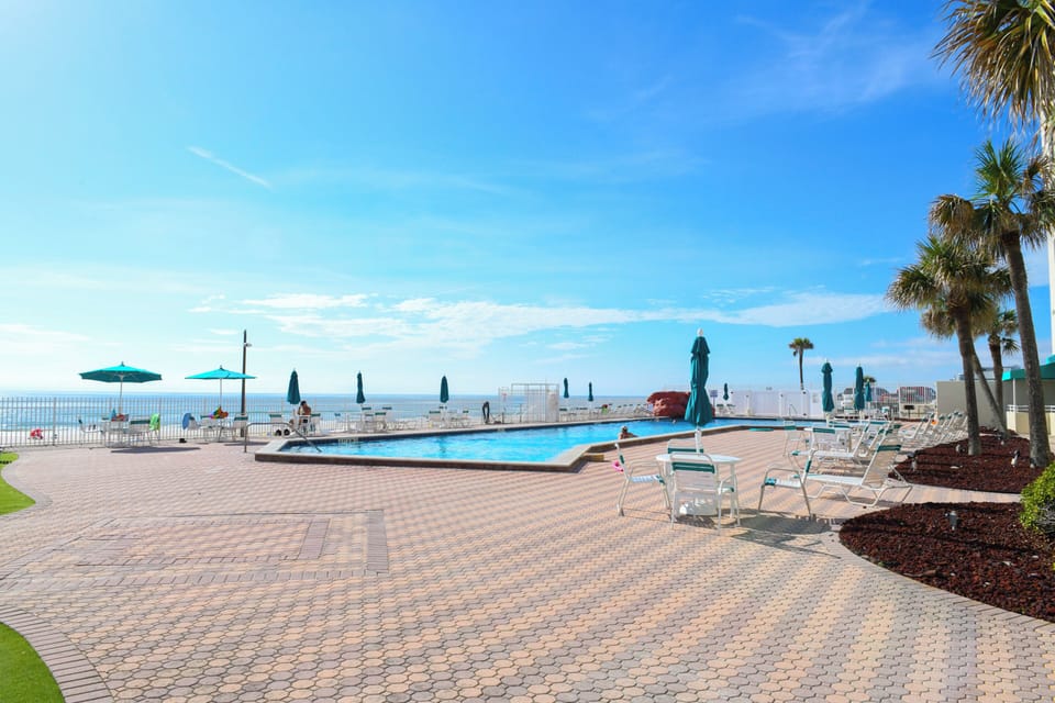 A spacious patio pool deck with palm trees borders a sparkling swimming pool overlooking the ocean. The property features beachfront access, sun loungers, and ample outdoor space for relaxation.