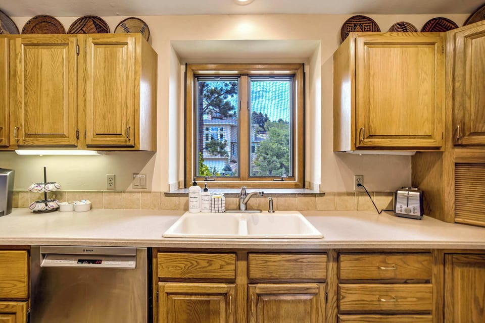A window above the sink frames leafy outdoor views, with white countertops and a stainless steel dishwasher rounding out this well-equipped prep space.
