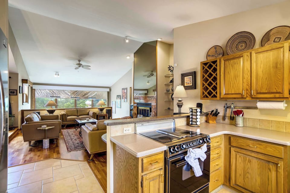 The kitchen opens onto the main living area, featuring warm oak cabinetry, white countertops, and a peninsula with seating that keeps the cook connected to the group.
