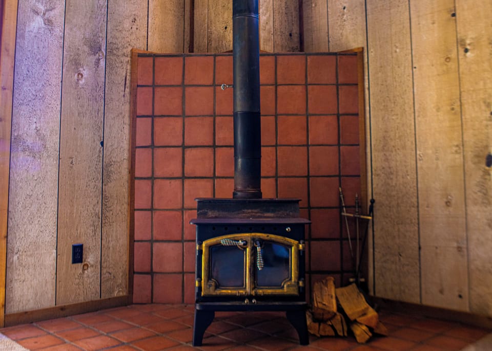 Your cozy wood-burning stove stands ready to warm your space on cool evenings, its golden-framed glass door glowing against rustic wood-paneled walls.