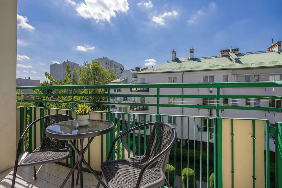 A balcony with outdoor seating and a small table. It overlooks nearby buildings and greenery.