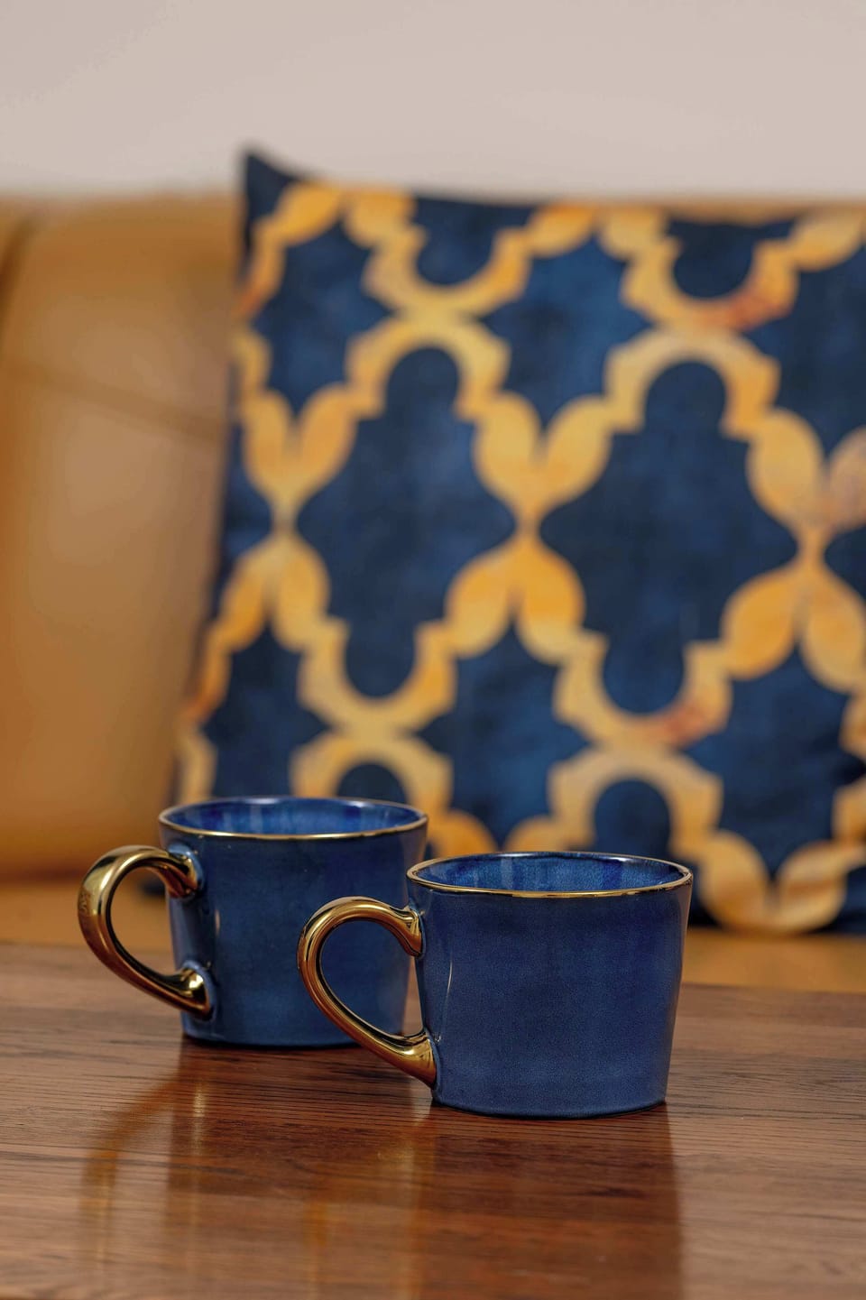 A close-up focus captures two dark blue ceramic mugs resting on a polished wooden surface. In the soft-focus background, a blue and gold geometric patterned pillow rests on a leather sofa.