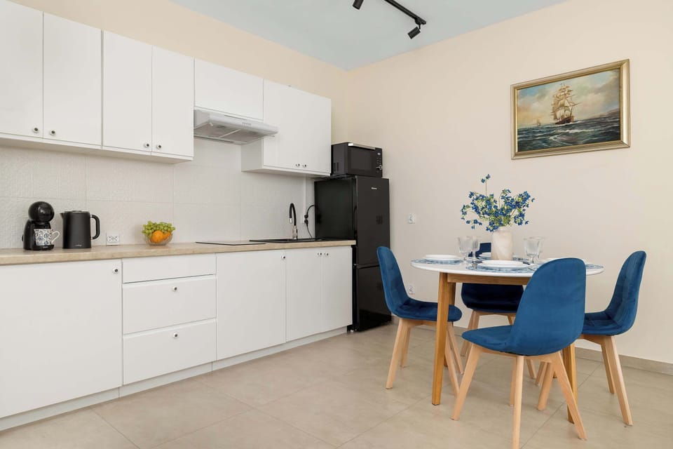A spacious open-concept kitchen area displays minimalist white cabinetry paired with a black refrigerator. A round blue dining table surrounded by matching plush velvet chairs provides a cozy spot for meals.