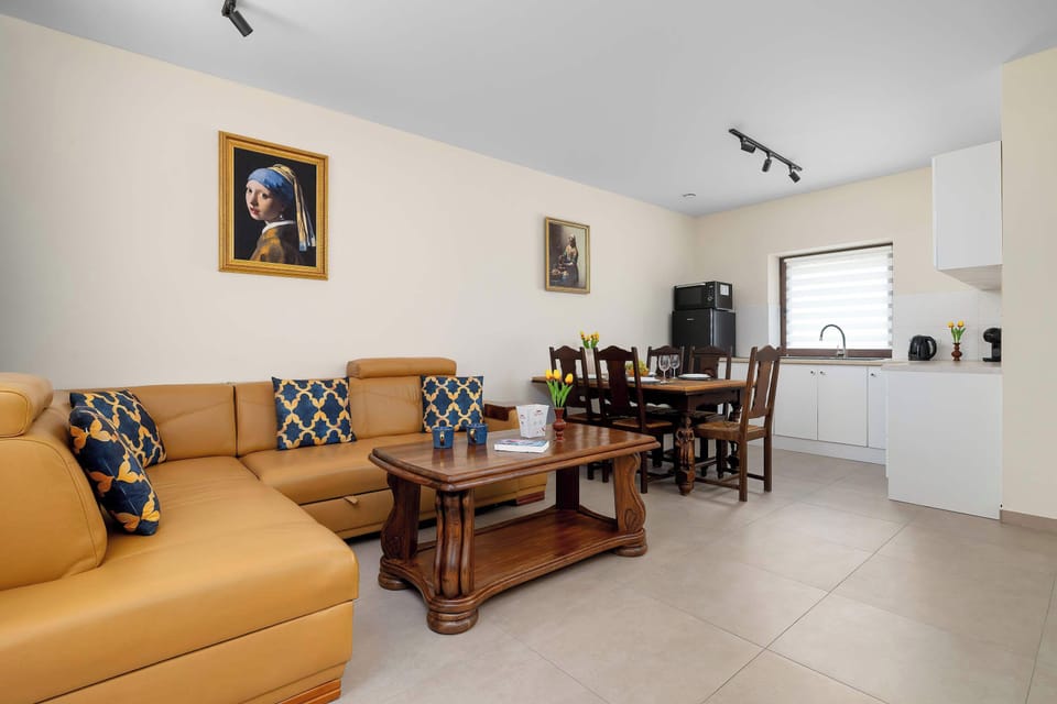 A generous living and dining area boasts a large L-shaped tan leather sofa arranged around a solid wood coffee table. In the background, a dark wooden dining table with high-backed chairs stands near the kitchen counter.