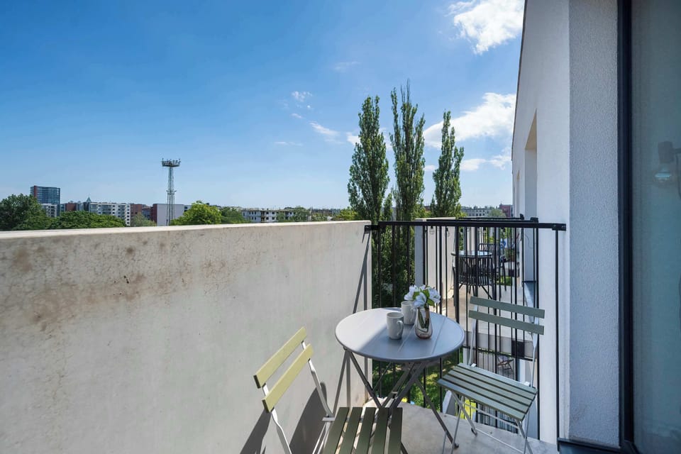 A view from the balcony reveals a sturdy metal railing and a compact outdoor table with chairs. Green treetops stretch across the horizon under a vast, bright blue sky with light clouds.