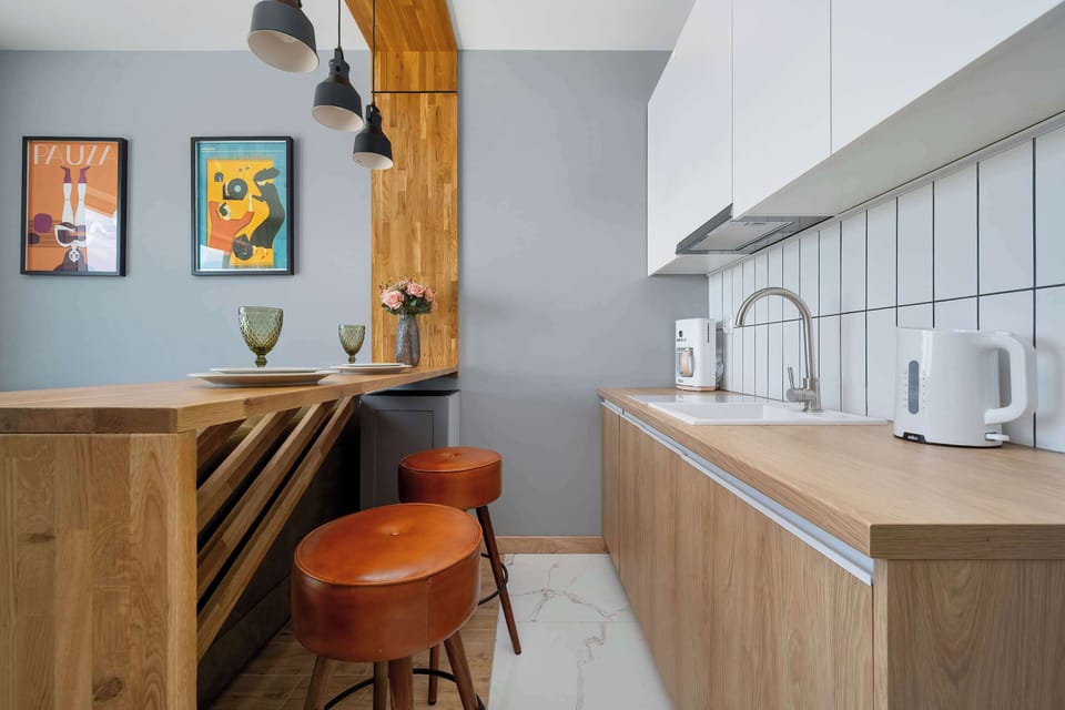 A functional wooden breakfast bar is paired with two elegant, cognac-colored leather bar stools. In the background, a minimalist kitchen corner showcases light wood lower cabinets, a clean white tiled backsplash, and a white electric kettle.