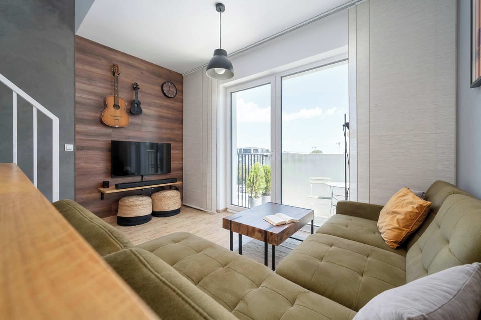 This wide angle of the living space highlights the open-concept layout connecting the seating area to the balcony. A flat-screen television is mounted on a rich wooden accent wall, complemented by woven wicker stools underneath.