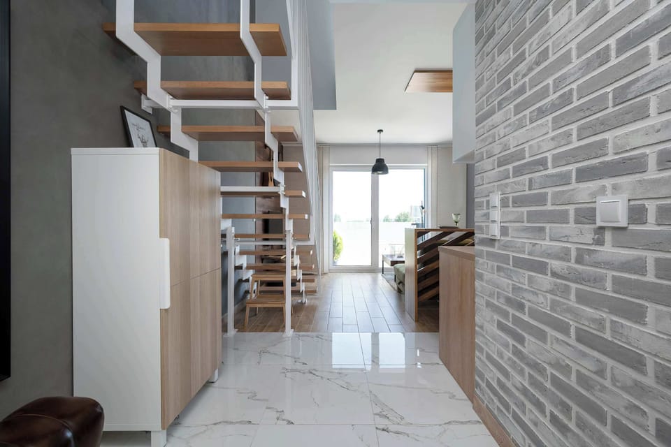The view from the entryway looks past a white storage cabinet and a wooden partition toward the bright living area. Polished white floor tiles reflect the natural light coming from the far window.