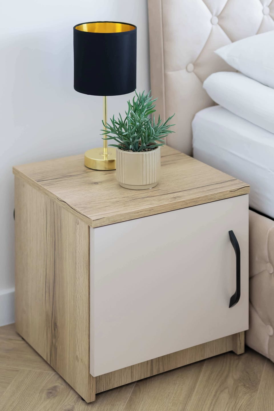 A detailed shot highlights a compact bedside table crafted from light wood with a dark metal handle. On its surface, a modern black and gold lamp sits next to a tiny, decorative potted succulent.