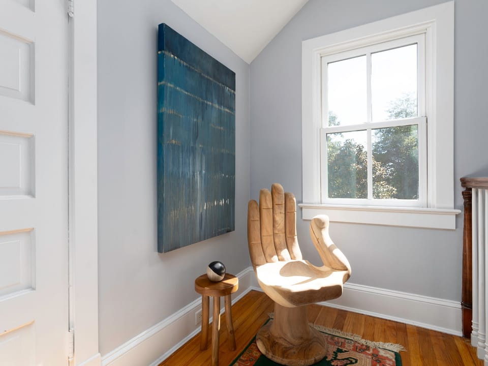 A sunlit nook features a unique hand-carved wooden chair, abstract art, and warm hardwood floors—your perfect spot to unwind and soak in the peaceful ambiance.