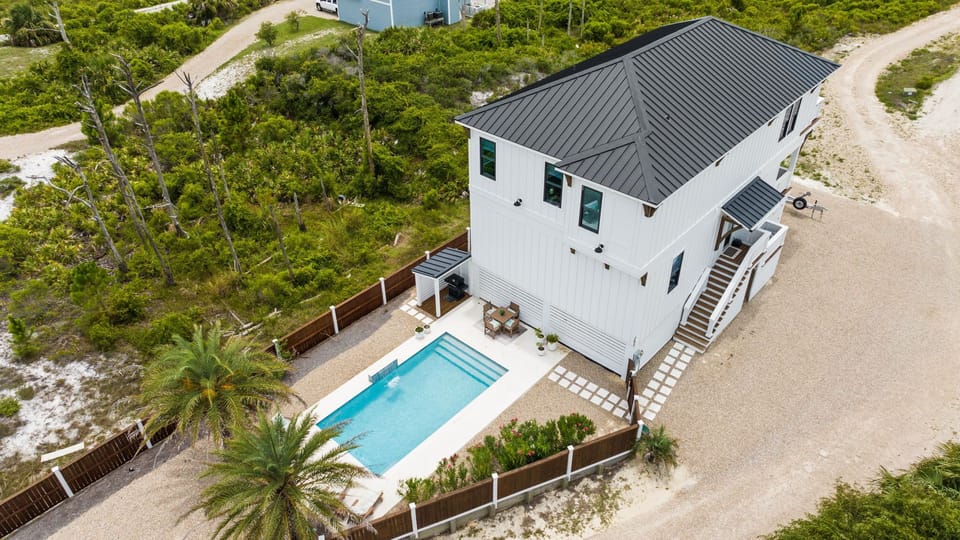Aerial view of a modern coastal vacation home surrounded by natural vegetation, featuring a private pool and convenient parking access along a sandy island road.