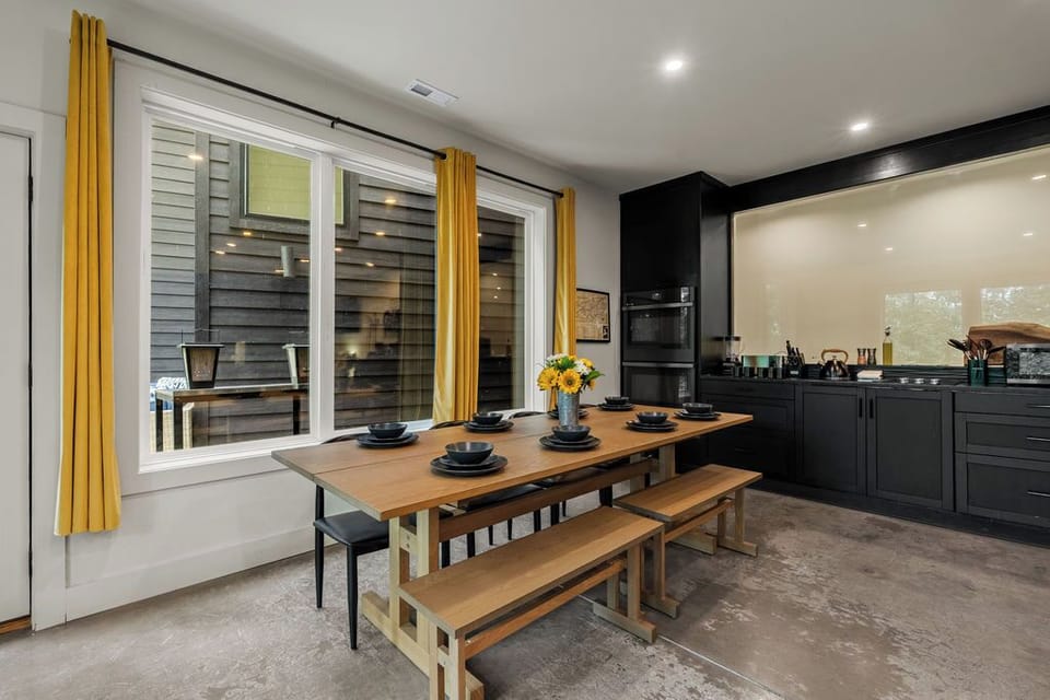 Open dining room with long table, bench seating, modern black kitchen, and large windows facing the forest.