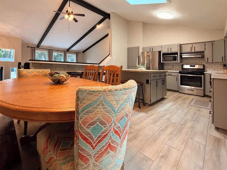 Open-plan dining and kitchen with round wood table, vaulted beamed ceiling, and stainless appliances — the natural gathering spot for the group