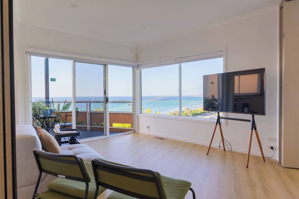 A roomy living area opens to a private balcony with sweeping 200-degree views of the Pacific Ocean, Phillip Bay, and Melbourne's skyline. Furnished with a comfy sofa and equipped with a Smart TV featuring YouTube/Netflix and HDMI connectivity.