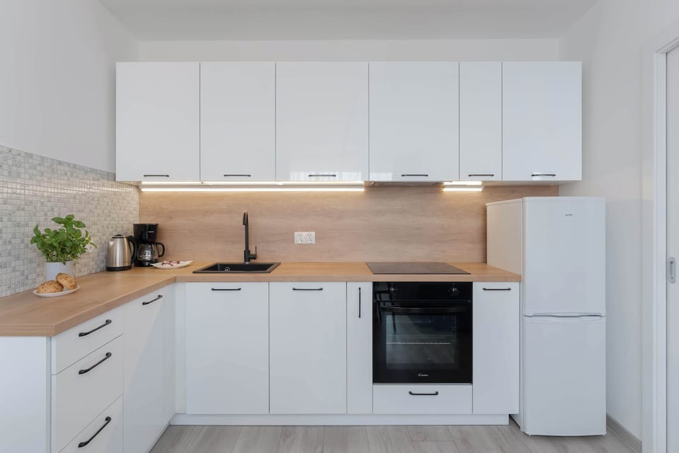 A sleek, contemporary kitchenette is equipped with minimalist white cabinetry and a warm wood-toned countertop. A freestanding white refrigerator sits neatly at the end of the counter line.