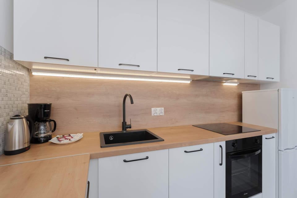 A close-up of the kitchenette workspace showcases a clean induction cooktop and a built-in black oven. Under-cabinet LED lighting illuminates the smooth wooden countertop and stainless steel sink.