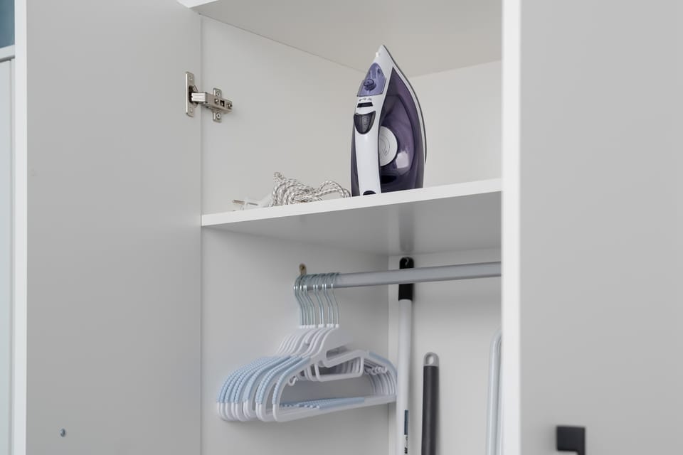 An open white wardrobe shelf holds a modern steam iron and several clothes hangers. This dedicated storage space keeps garments organized and wrinkle-free.