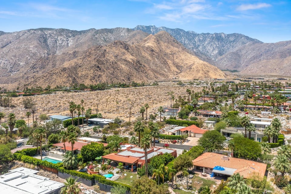 Aerial view of the property nestled in a desert landscape, with dramatic mountain ranges rising in the background and palm-lined neighborhoods dotting the valley below.