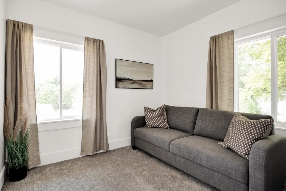 Gray fabric sofa with patterned pillows sits between two windows framed by beige curtains in a carpeted room.