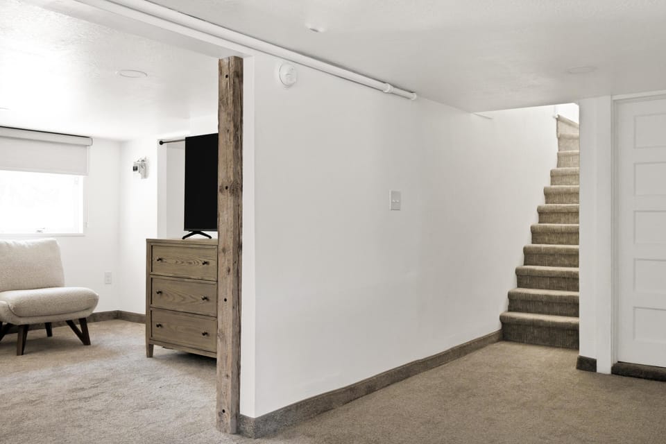 Carpeted stairway leads upstairs beside a white wall and wooden post, with a beige armchair and dresser in adjacent room.