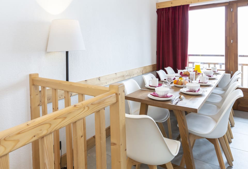 The dining area set for ten, with natural light pouring through the red-curtained windows and the railing of the upper level visible beyond.