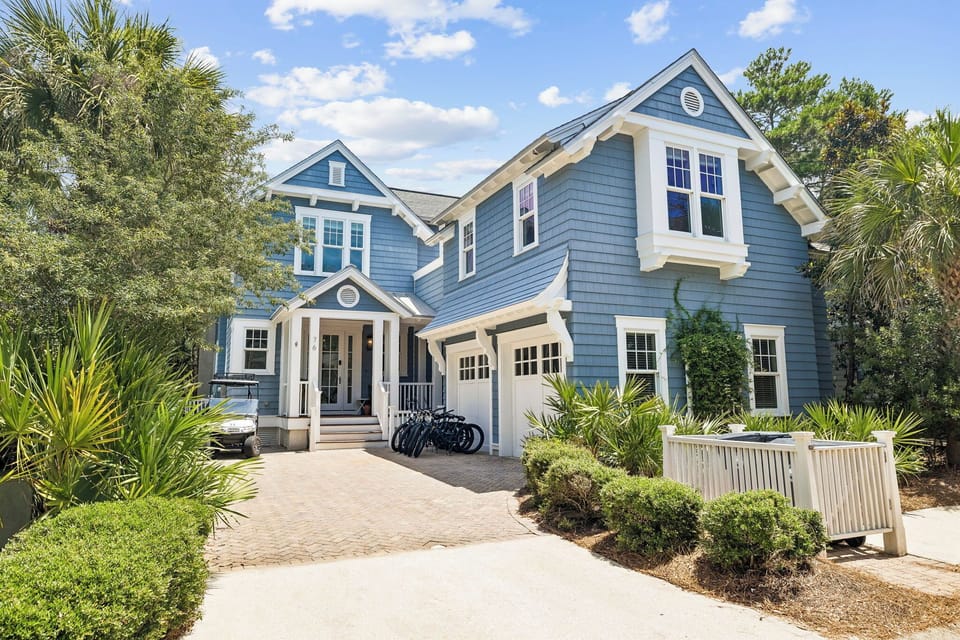 Charming coastal-style home with blue siding, white trim, and welcoming front porch, surrounded by lush tropical landscaping and palms.