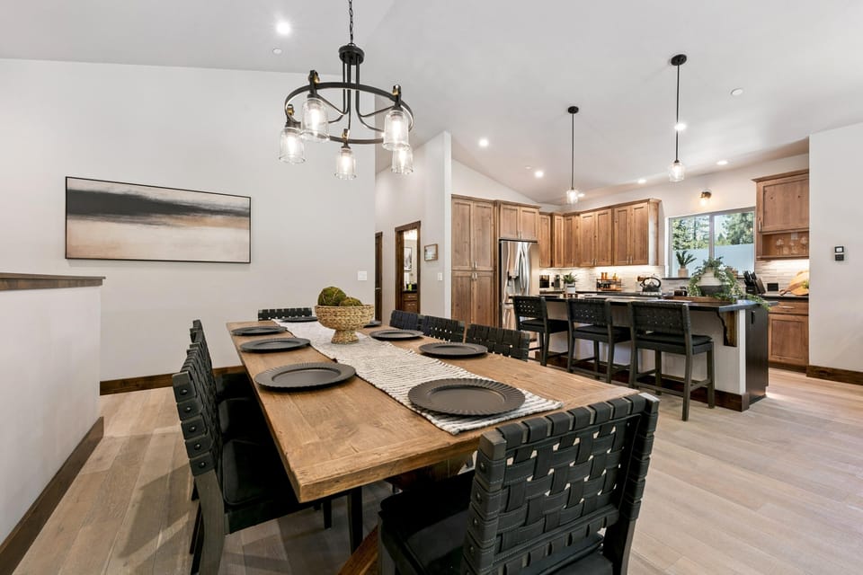 Black leather strap chairs surround a solid wood table set with black plates beneath a glass jar chandelier.