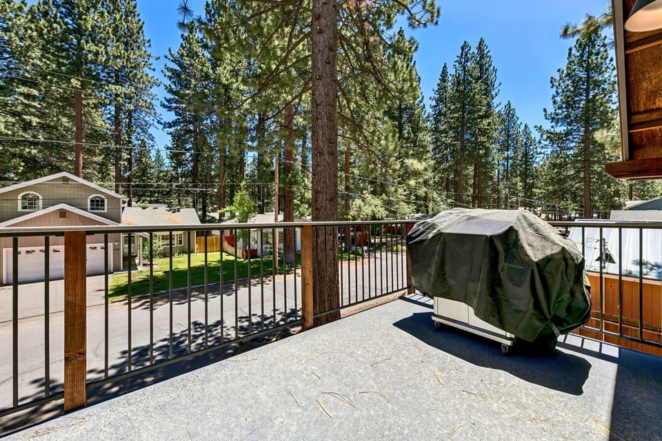 Second-floor balcony with black metal and wood railing, covered grill, and views of tall pine trees.