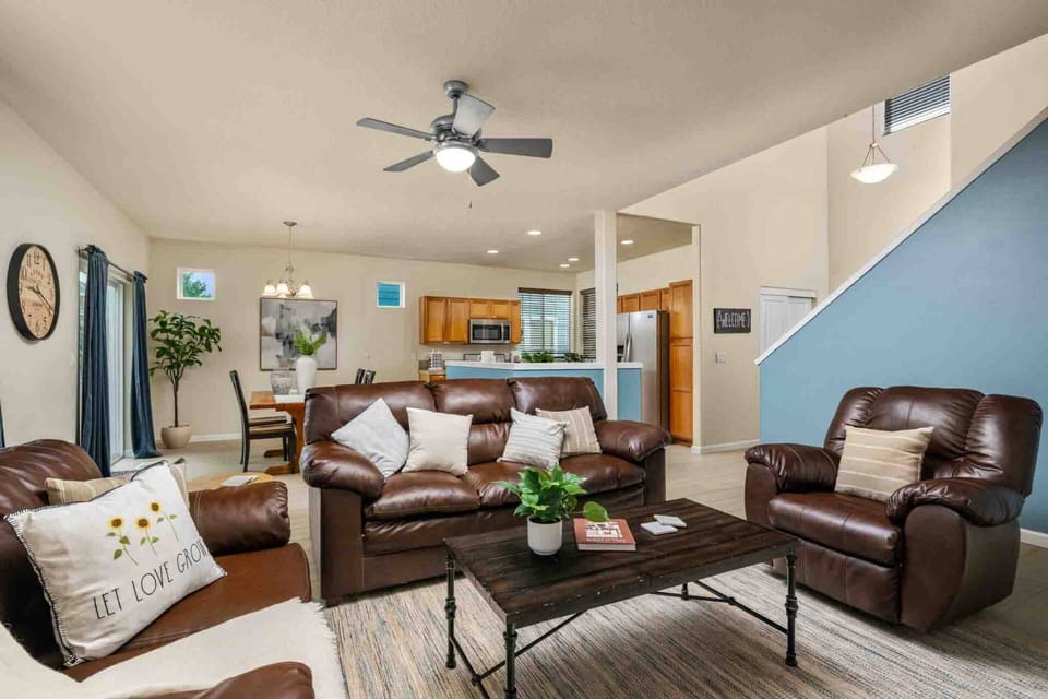 A broad view of the open main level, showcasing comfy leather sofas, a ceiling fan, light wood-look floors, and open sightlines to the dining room and