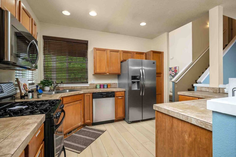 A functional kitchen complete with warm wooden cabinetry, stainless steel appliances, tile countertops, and direct view toward the main entryway stair