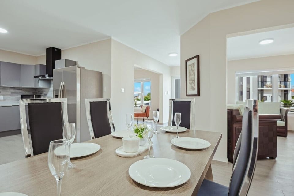 The open dining area features a light wood table with seating for guests, connecting the modern kitchen to the bright, inviting living room.
