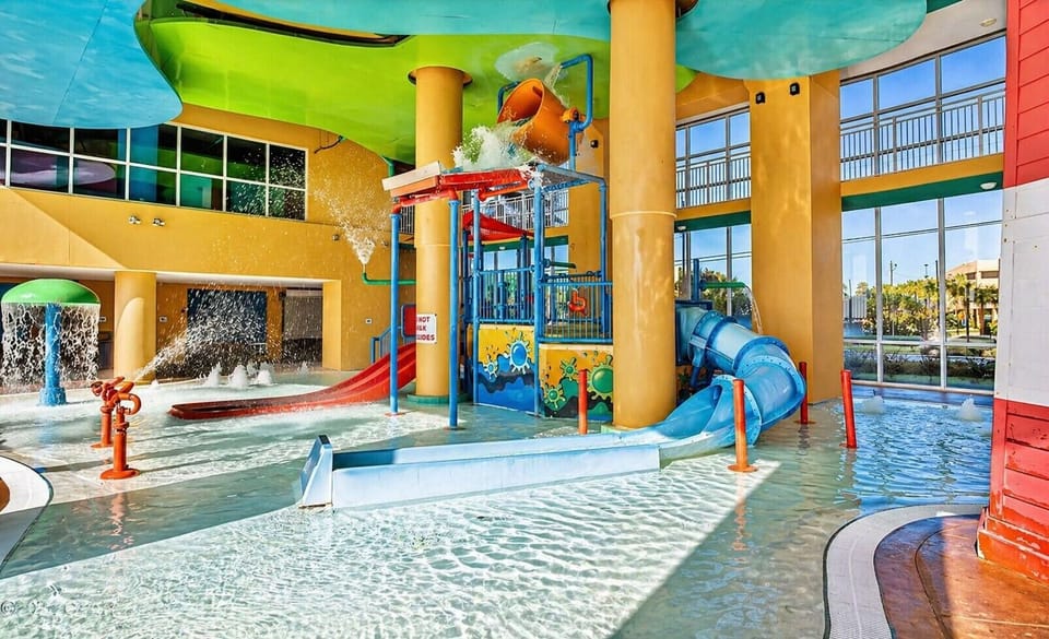Vibrant indoor aqua park featuring colorful slides, splash zones, and a tipping bucket for endless family fun under a sun-soaked glass ceiling.
