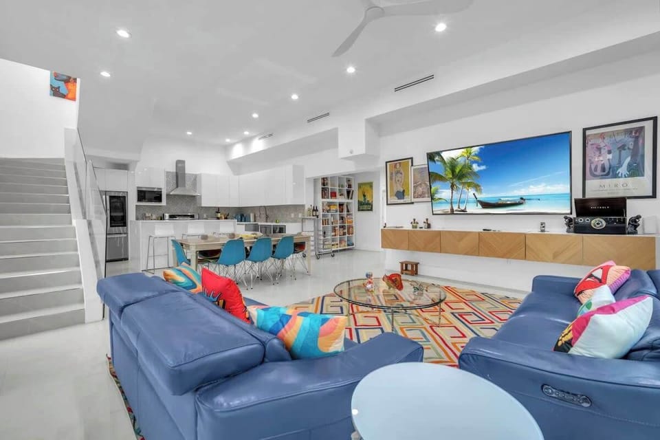 Comfortable family living room featuring a large-screen TV, oversized blue sectional, and a picture-perfect backdrop of tropical greenery and pool.