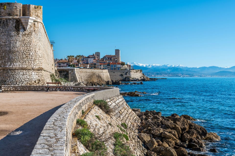 Stone fortification walls curving along the Antibes coastline with snow-capped mountains visible across the bay