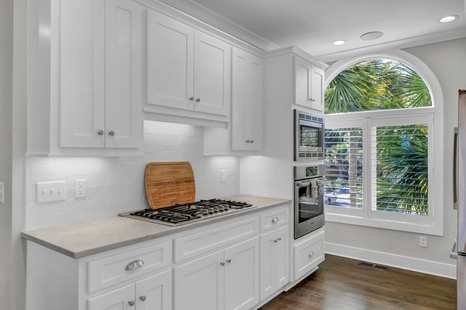 Your bright, modern kitchen features sleek white cabinetry, stainless-steel appliances, and an arched window framing lush tropical greenery—perfect for preparing meals in natural light.