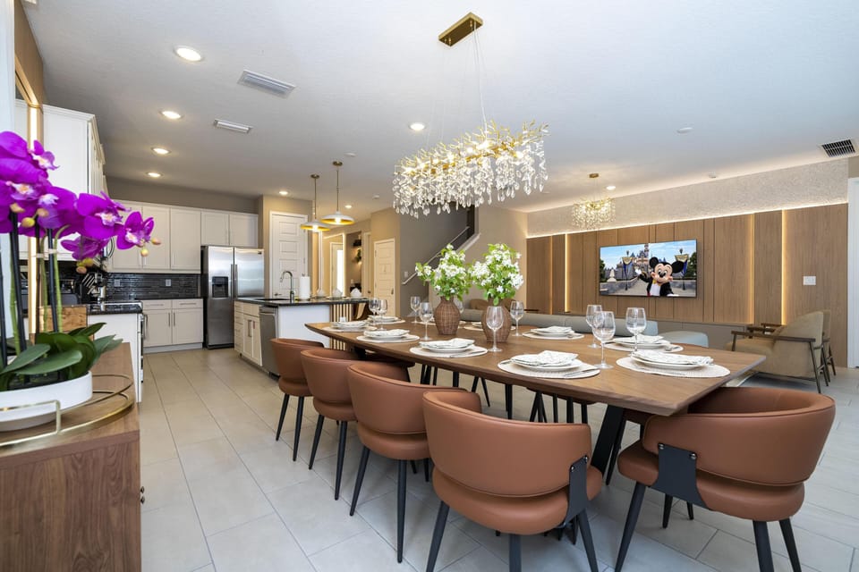 Long wood dining table set with white plates, surrounded by brown chairs under a crystal chandelier.