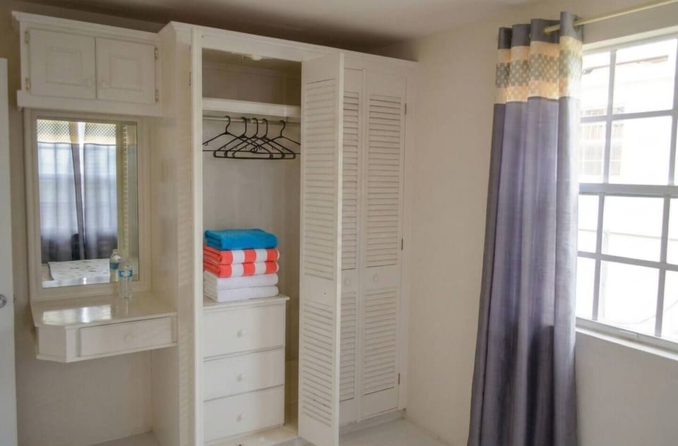 Bright, clean room featuring a built-in closet, hangers and drawers, a handy vanity setup with a mirror, and fresh linens. A large window brings in beautiful natural light, framed by stylish privacy curtains. Perfect, organized space for your stay!