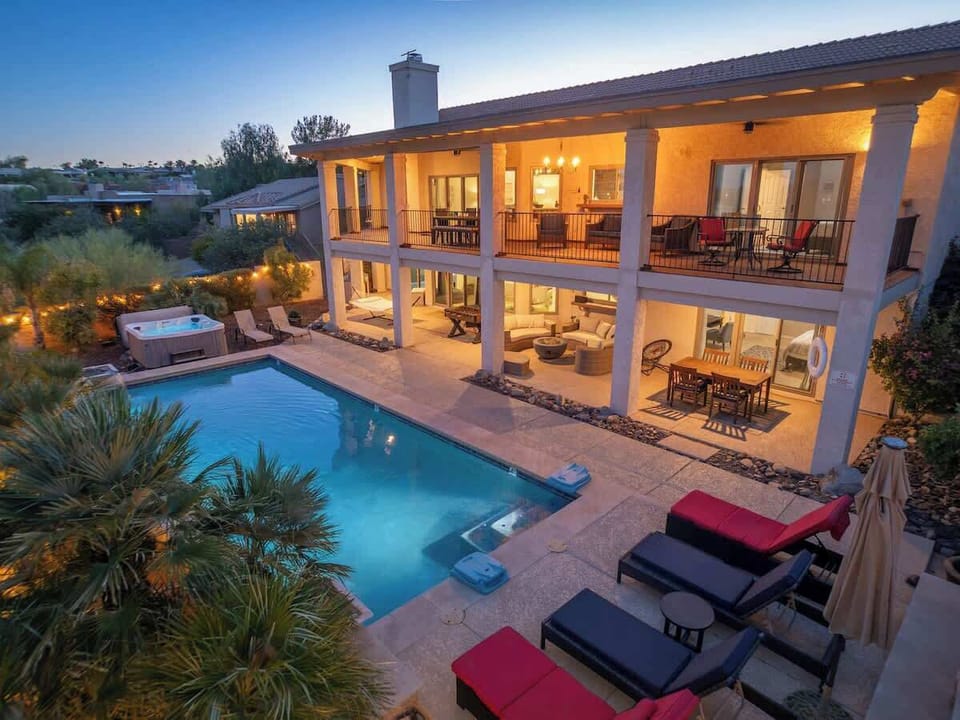 This stunning two-story retreat showcases a sparkling pool and hot tub framed by elegant outdoor living spaces, captured during golden hour when warm interior lighting creates an inviting glow throughout.