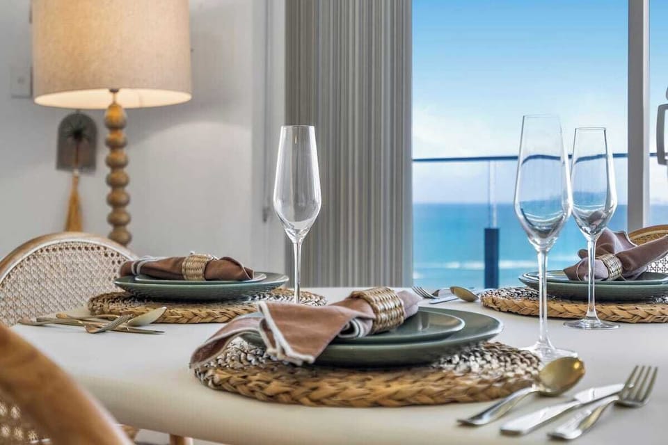 Scenic Dining Detail An elegant tablescape complete with woven placemats, polished cutlery, and crystal glassware. Overlooking the bright blue ocean, it offers a sophisticated backdrop perfect for sharing a coastal vacation meal.