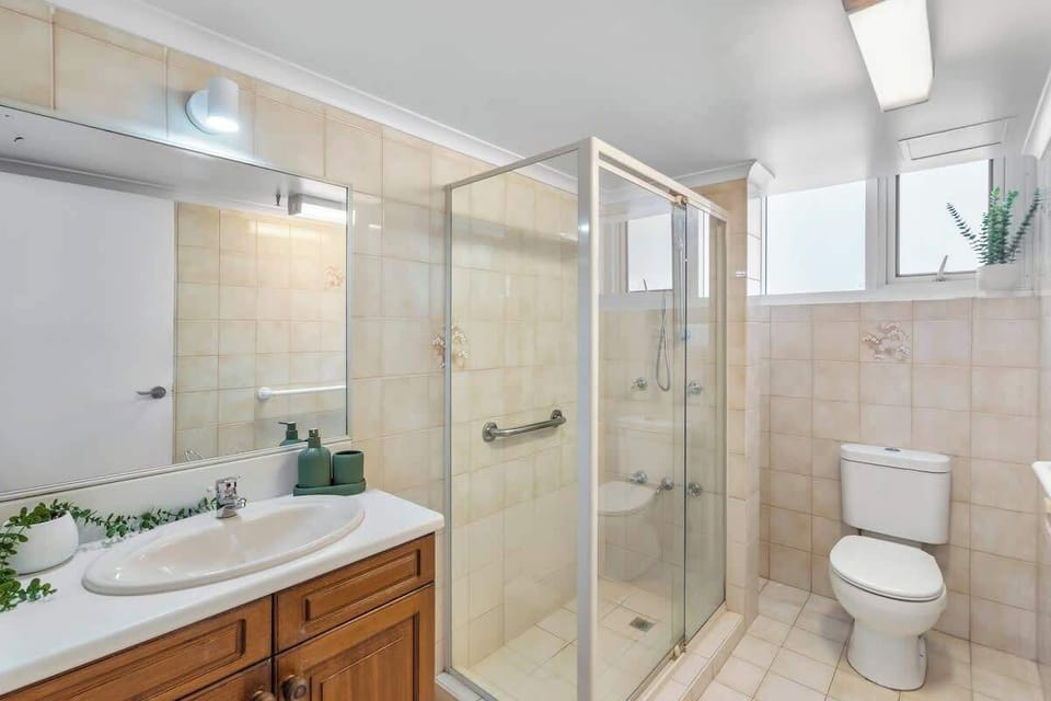 Unwind in this spacious secondary bathroom, offering full wall-to-wall tiling, a glass-enclosed walk-in shower, a wide vanity with wooden cabinetry, a large wall mirror, and modern fixtures.