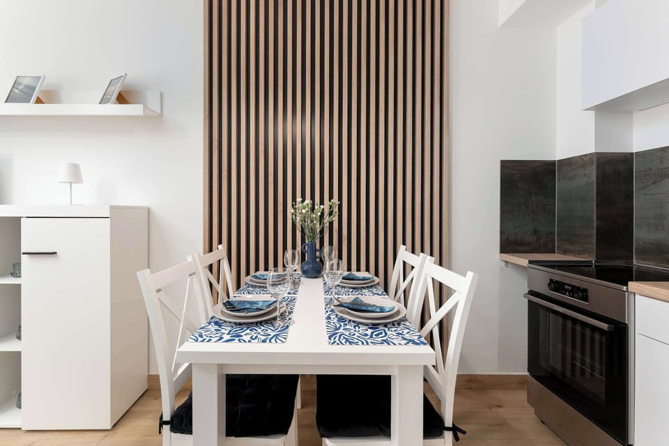 A dining table neatly set for four sits adjacent to a built-in oven and black induction cooktop. A vertical wooden slat partition creates a stylish divider.