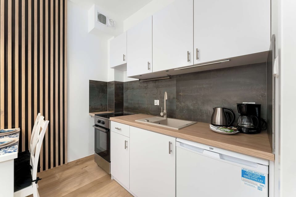 A compact kitchenette displays clean white cabinets and a warm wood-toned countertop. A black electric kettle and coffee maker sit near a stainless steel sink.