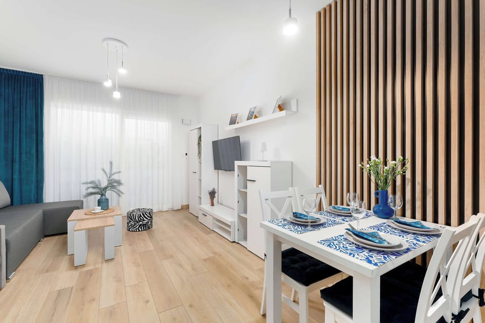 A white wooden dining table with four chairs sits in front of a decorative vertical wood slat wall feature. A blue vase with flowers serves as a centerpiece.