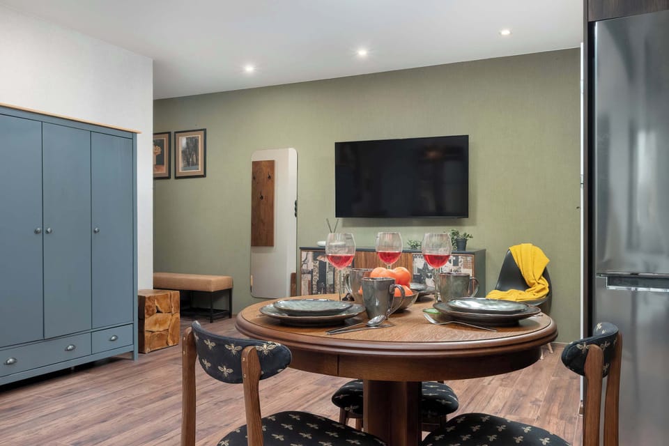 A round wooden dining table surrounded by matching chairs sits prominently in front of an olive green accent wall. A flat-screen television is mounted above the table, positioned next to a tall grey storage wardrobe.