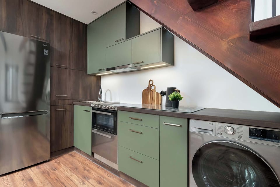 The kitchen setup under the stairs is efficiently organized with a washing machine integrated into the green cabinetry. A stainless steel stove and oven unit sit beneath a minimalist extractor hood.