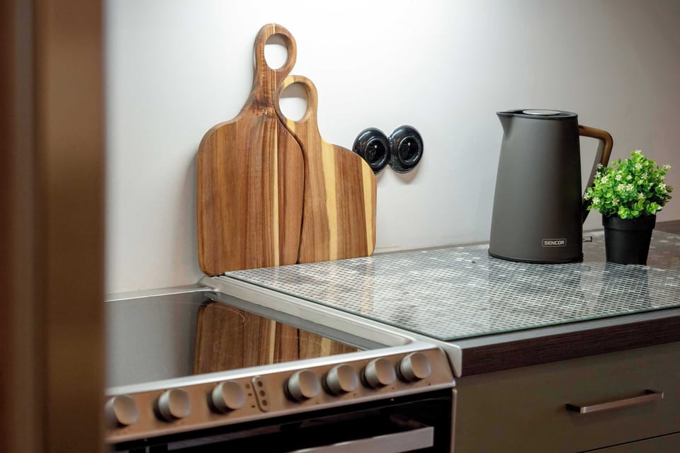 Wooden cutting boards stand upright against the wall on a textured granite kitchen countertop. A modern black electric kettle rests nearby next to a small potted plant.